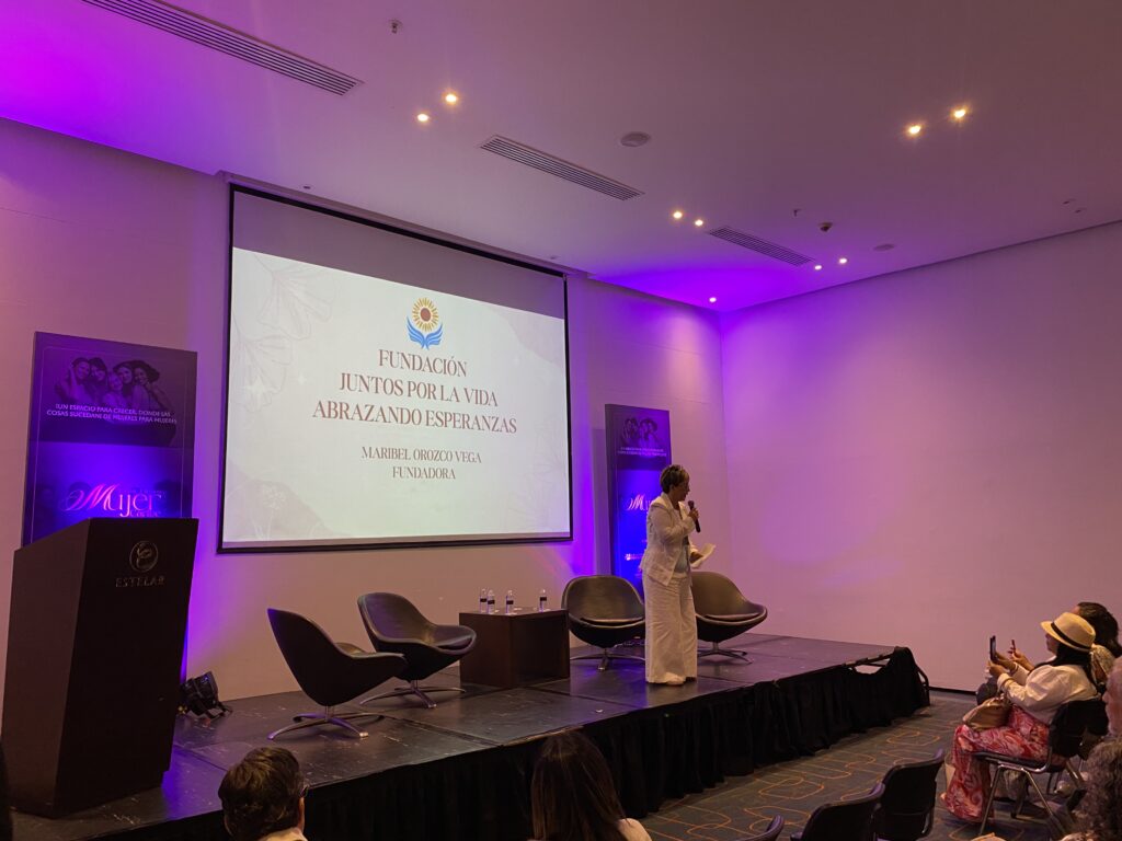 La fundadora expone en el escenario de Expo Mujer, iluminado en morado, frente a la pantalla titulada «Fundación Juntos por la Vida: abrazando esperanzas».