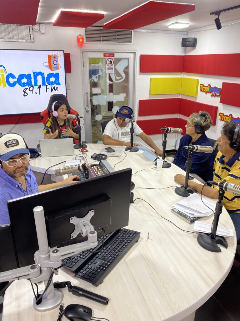 La cabina de Tropicana 89.1 FM durante una emisión, con el equipo sentado a la mesa redonda con audífonos y micrófonos.