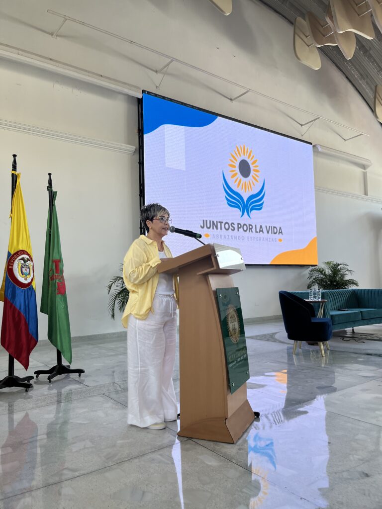 La Dra. Maribel Orozco habla desde el atril, junto a la bandera de Colombia y la pantalla con el logo de Juntos por la Vida.