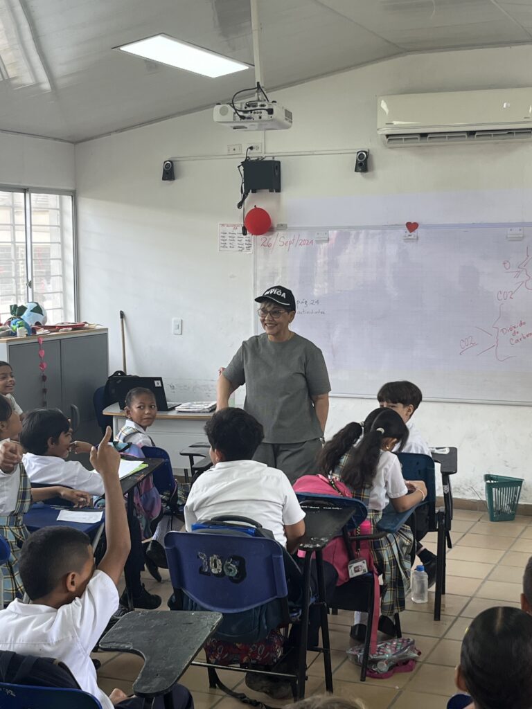 La Dra. Maribel Orozco, con gorra de la Policía Cívica, conversa con niños de uniforme escolar sentados en sus pupitres; un niño levanta la mano.
