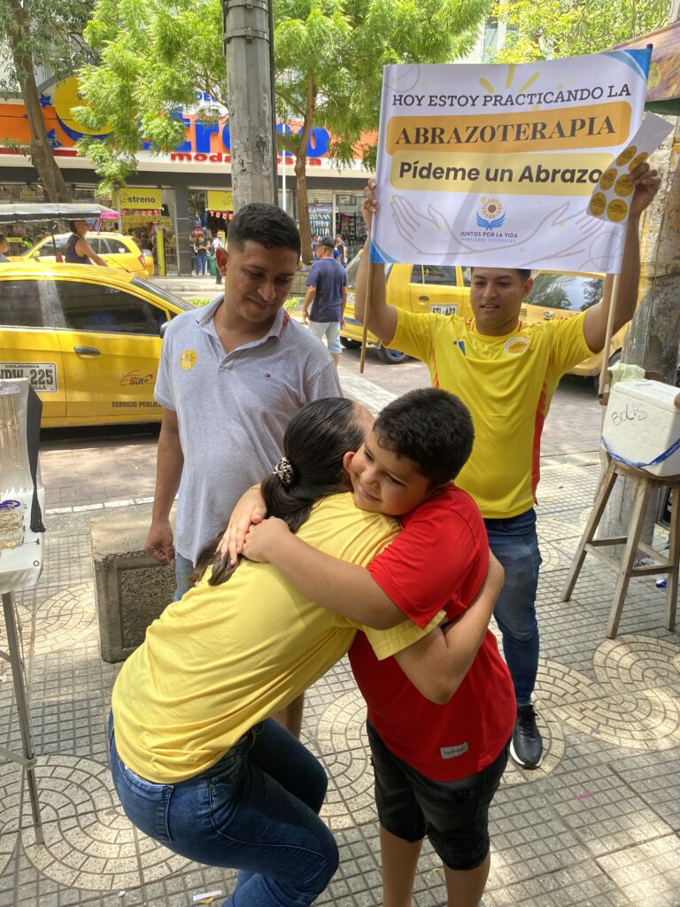 Una voluntaria de camiseta amarilla abraza a un niño en plena calle del Paseo Bolívar, bajo un cartel que dice «Hoy estoy practicando la abrazoterapia: pídeme un abrazo».