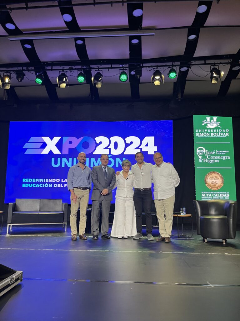Cinco panelistas posan en el escenario de ExpoUnisimón 2024, frente a la pantalla azul del evento y el pendón de la Universidad Simón Bolívar.