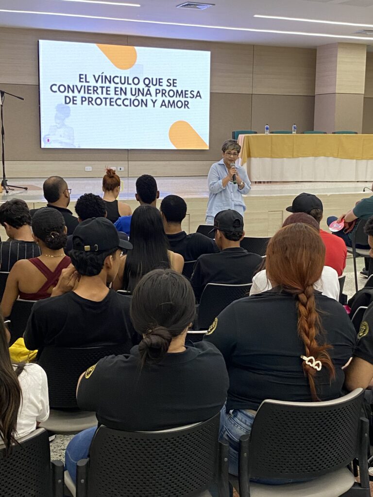 La Dra. Maribel Orozco habla ante estudiantes sentados de espaldas, frente a una diapositiva que dice «El vínculo que se convierte en una promesa de protección y amor».