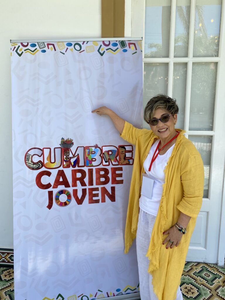 La Dra. Maribel Orozco, de amarillo, junto al pendón de la Cumbre Caribe Joven.