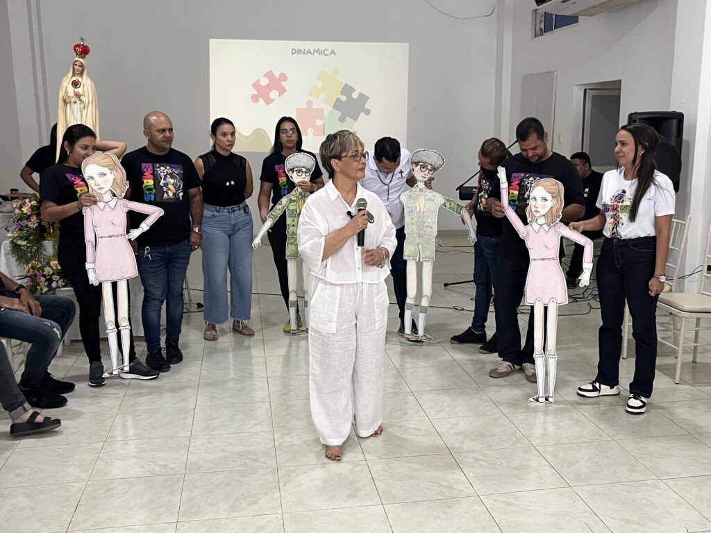 La Dra. Maribel Orozco habla con micrófono en el salón de la comunidad de Emaús, mientras los participantes sostienen siluetas de cartón durante la dinámica.