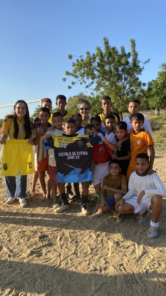 Niños de la escuela de fútbol Juan 23 sostienen su nuevo uniforme en una cancha de arena, acompañados por la Dra. Maribel Orozco.